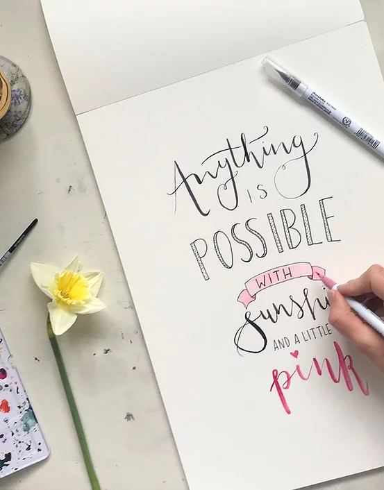 A hand doing calligraphy on a notepad "Anything is possible with sunshine and a little pink"