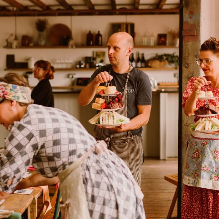 two women dressed in vintage style clothing and a man dressed in a chef's apron serving afternoon tea