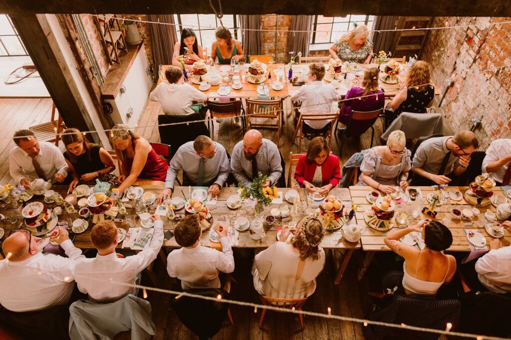 two long rustic tables laid for afternoon tea with guests sitting on either side of the table to celebrate a wedding